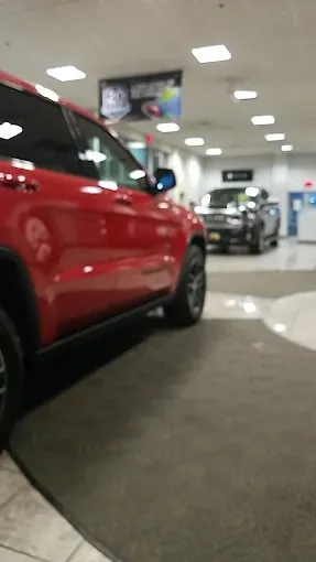 Red vehicle beside a floor mat inside a dealership showroom, with another vehicle in the background